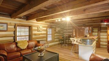 A log cabin interior with a leather sofa, a coffee table, a dining area with chairs, and a kitchen area with a sink and counter.