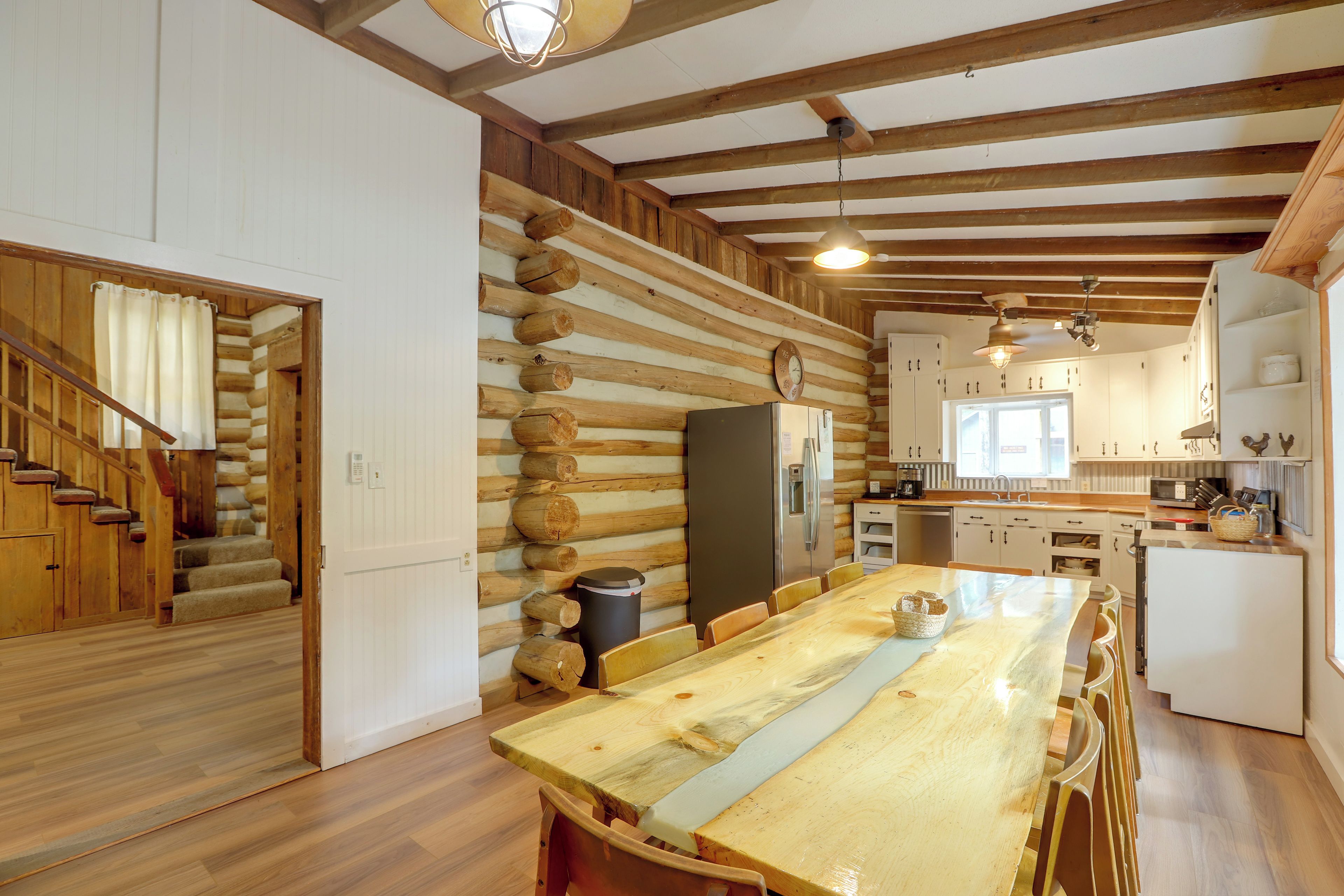 A log cabin interior with a wooden dining table, kitchen area, and a staircase leading to another room.