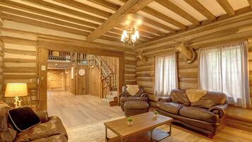 A log cabin interior with a fireplace, leather sofas, a dining table, and a staircase leading to an upper level.