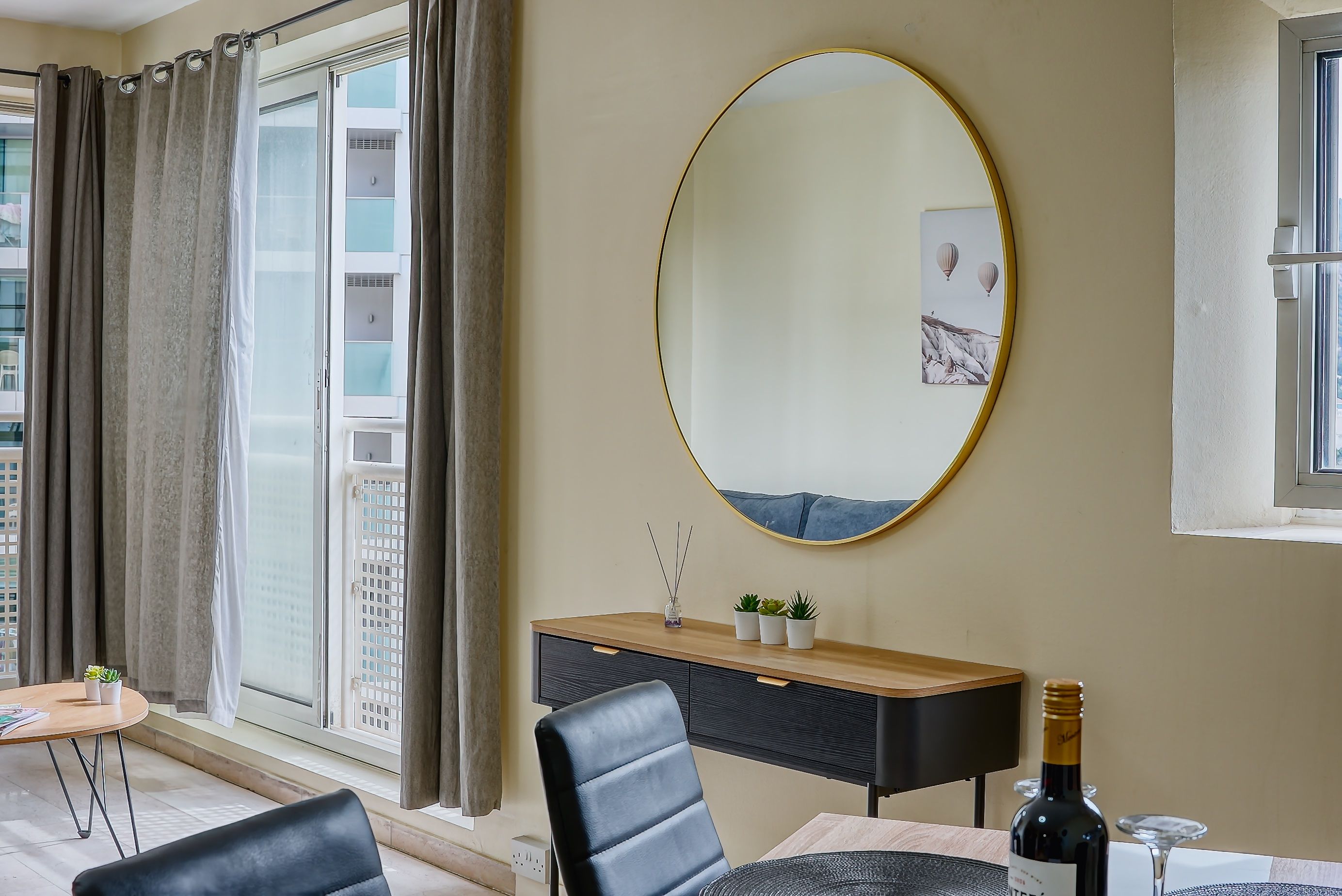 Una sala de estar moderna con un espejo ovalado grande, una consola de madera con objetos decorativos, una mesa de comedor con una botella de vino y vistas a un paisaje urbano a través de un ventanal amplio.