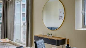 Una sala de estar moderna con un espejo ovalado grande, una consola de madera con objetos decorativos, una mesa de comedor con una botella de vino y vistas a un paisaje urbano a través de un ventanal amplio.