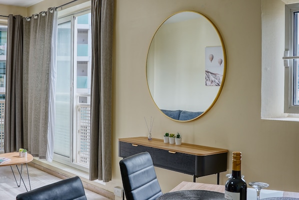 A modern living room with a large oval mirror, a wooden console table with decorative items, a dining table with a bottle of wine, and a view of a cityscape through a large window. - Eurotowers (Gibraltar)
