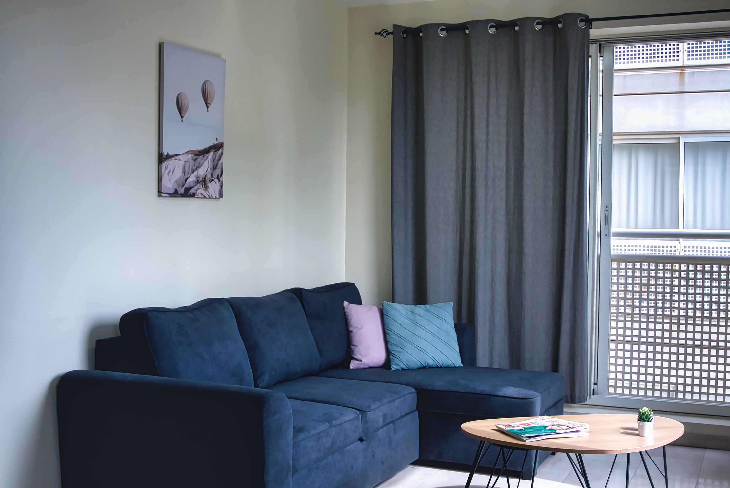 Una sala moderna con un sofá azul oscuro, una mesita redonda con una revista y un ventanal con vista a un edificio.