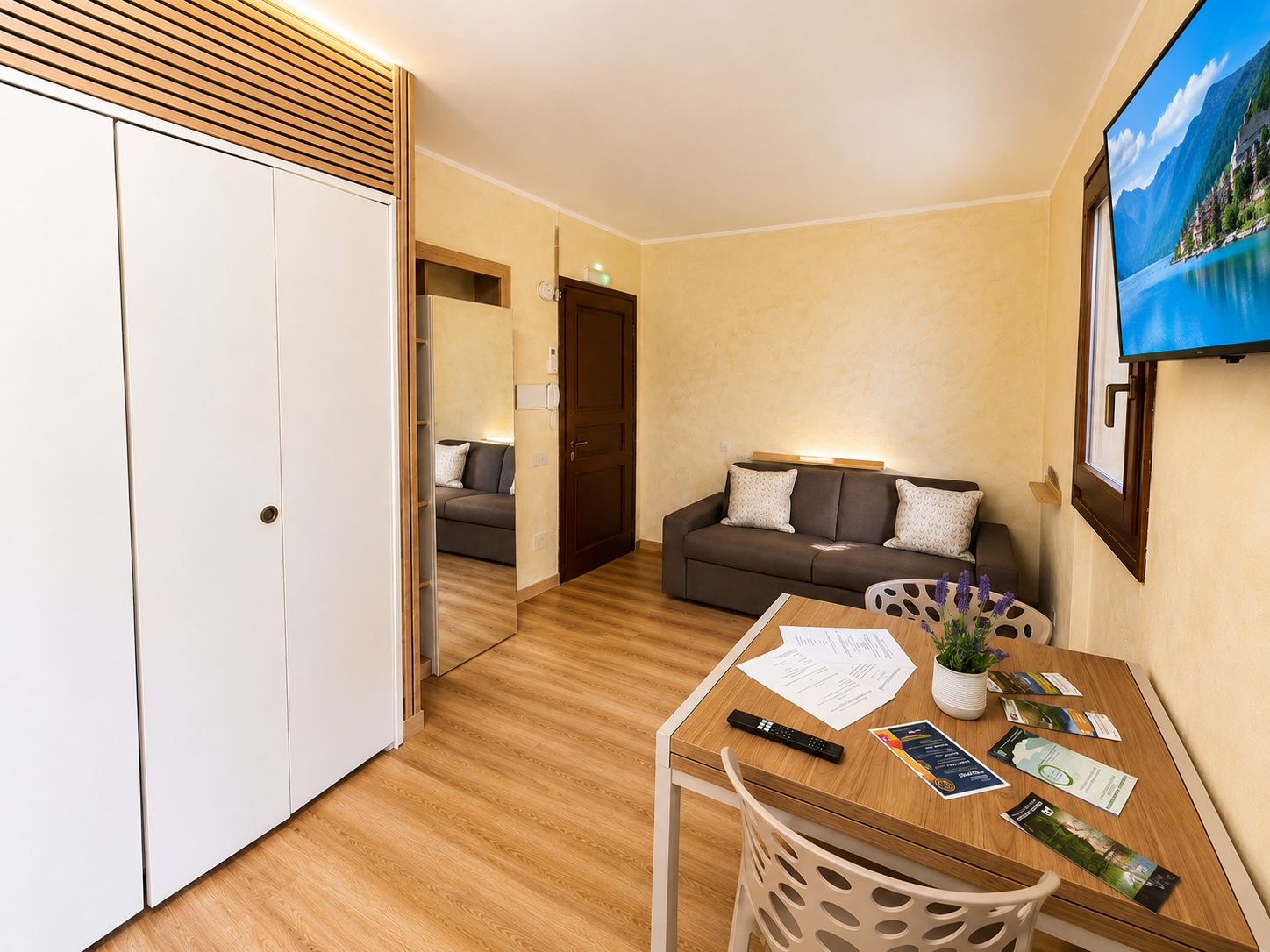 Ein Hotelzimmer mit Holzboden, einer Couch, einem Esstisch mit Papieren und einer Fernbedienung sowie einem Bild an der Wand.