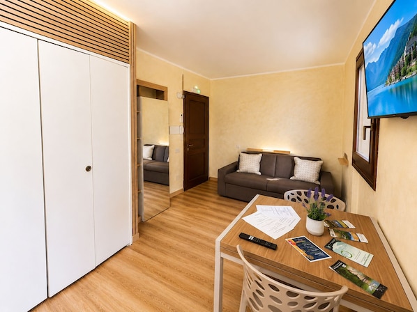 A hotel room with a wooden floor, a sofa, a dining table with papers and a remote control, and a painting on the wall. - Apartment with a balcony offering mountain views and a pool in a private residence (Introbio)