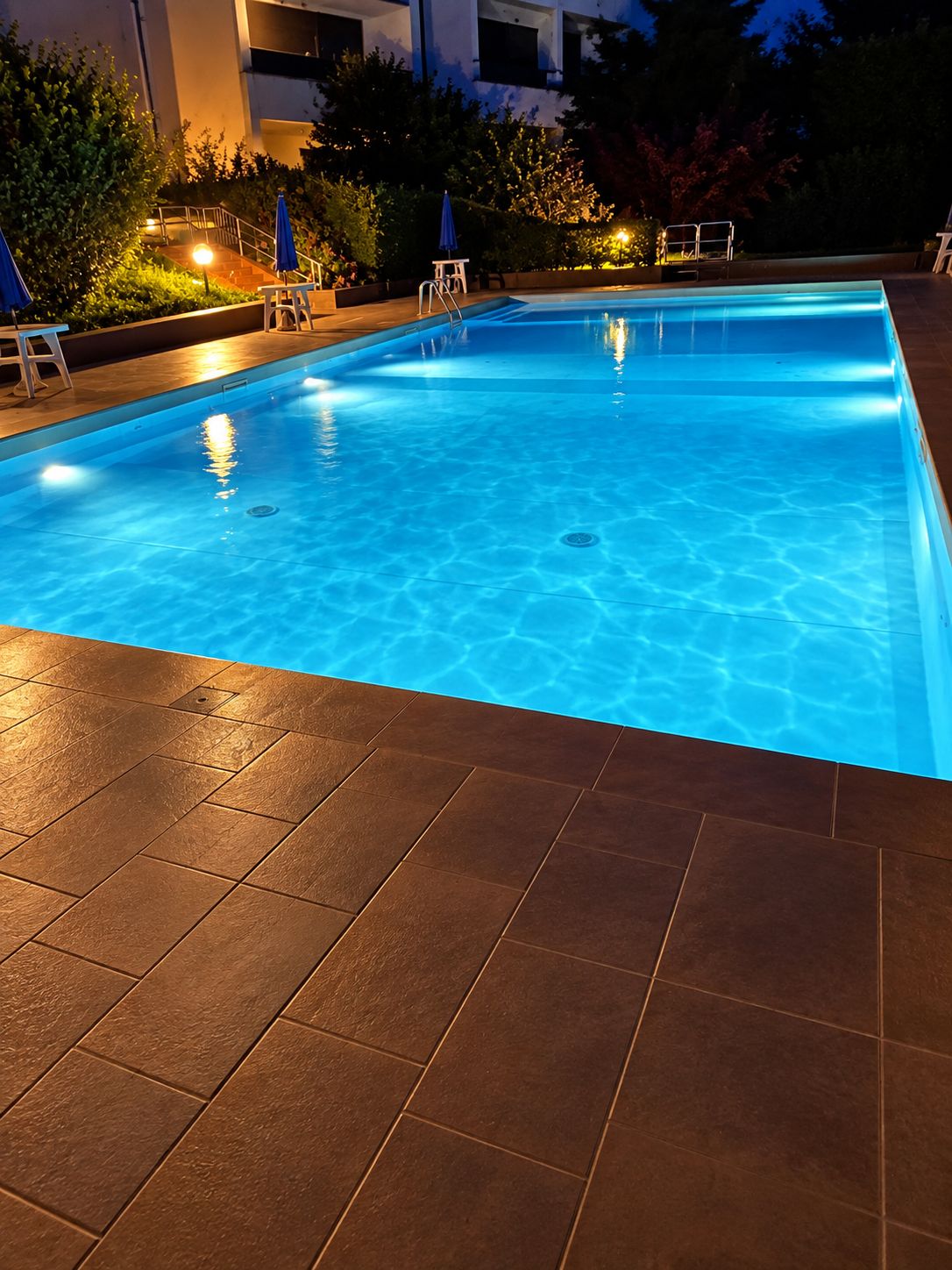 Ein Schwimmbecken mit Fliesen umgeben von Pflanzen und Gartenmöbeln.