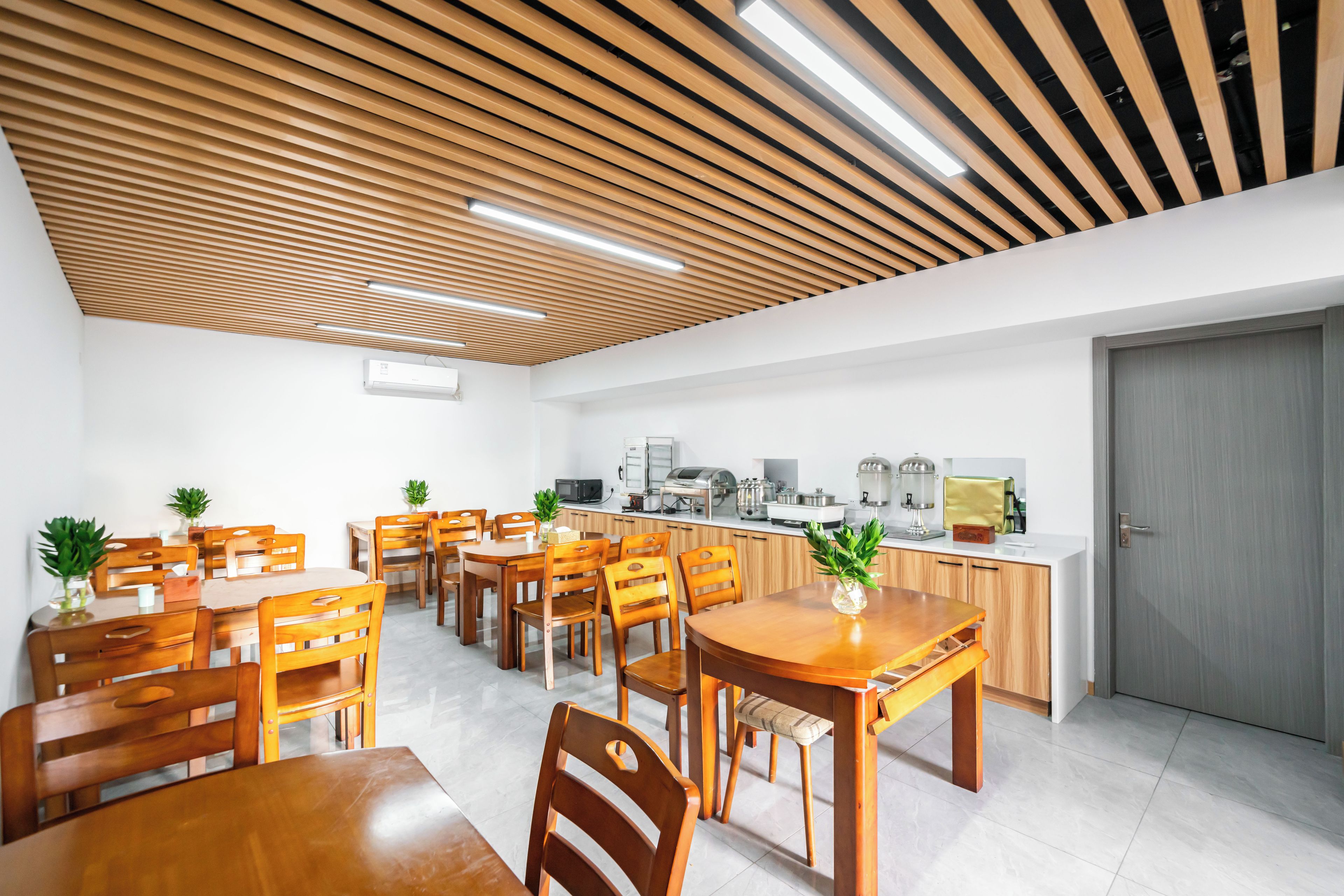 Ein moderner Essbereich mit Holztischen und -stühlen, eine Küche im Hintergrund und eine Decke mit Holzleisten und eingelassener Beleuchtung.