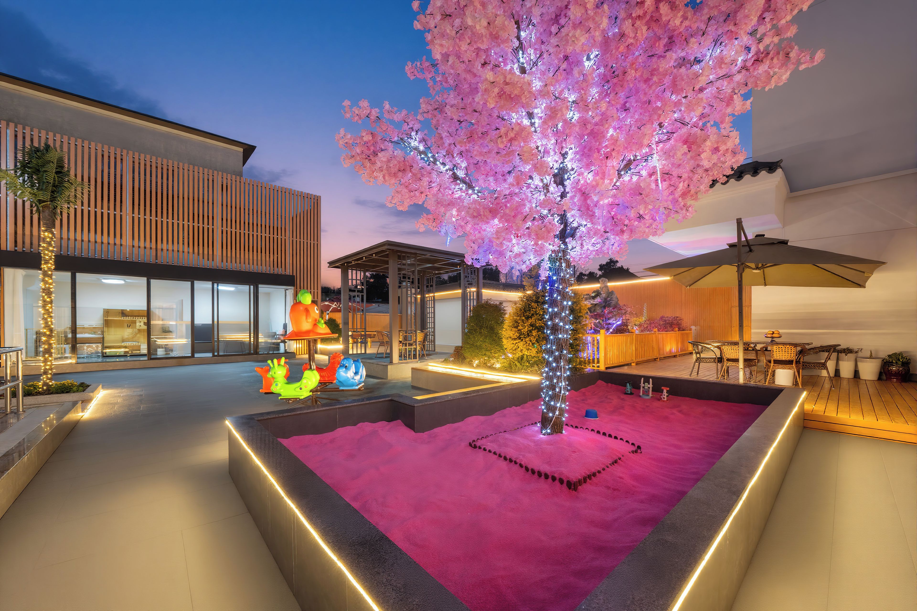 Un espacio exterior moderno con un árbol iluminado en rosa, asientos y un edificio con grandes ventanales.