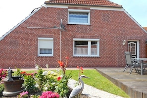 Ein Backsteinhaus mit geneigtem Dach, ein Garten mit Blumen und einem Vogel sowie eine Terrasse mit Gartenmöbeln.