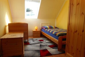 Ein kleines, gemütliches Schlafzimmer mit schrägen Wänden, einem Einzelbett, einem Nachttisch mit Lampe und einer hölzernen Kommode.