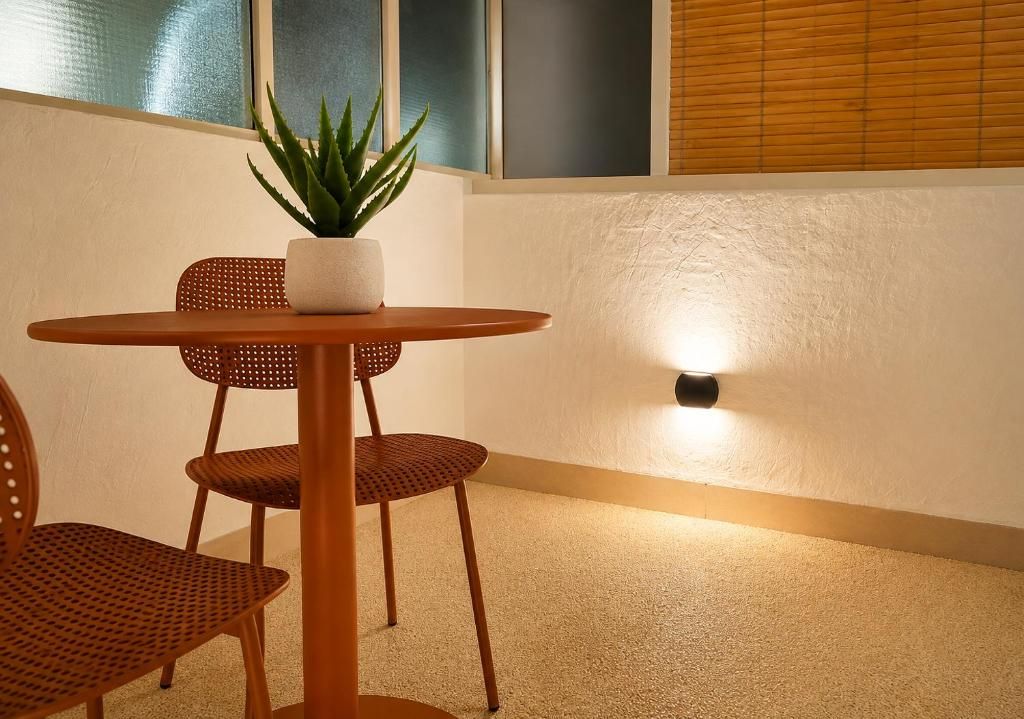Una pequeña mesa redonda con una planta en maceta, dos sillas de mimbre y una lámpara de pared.