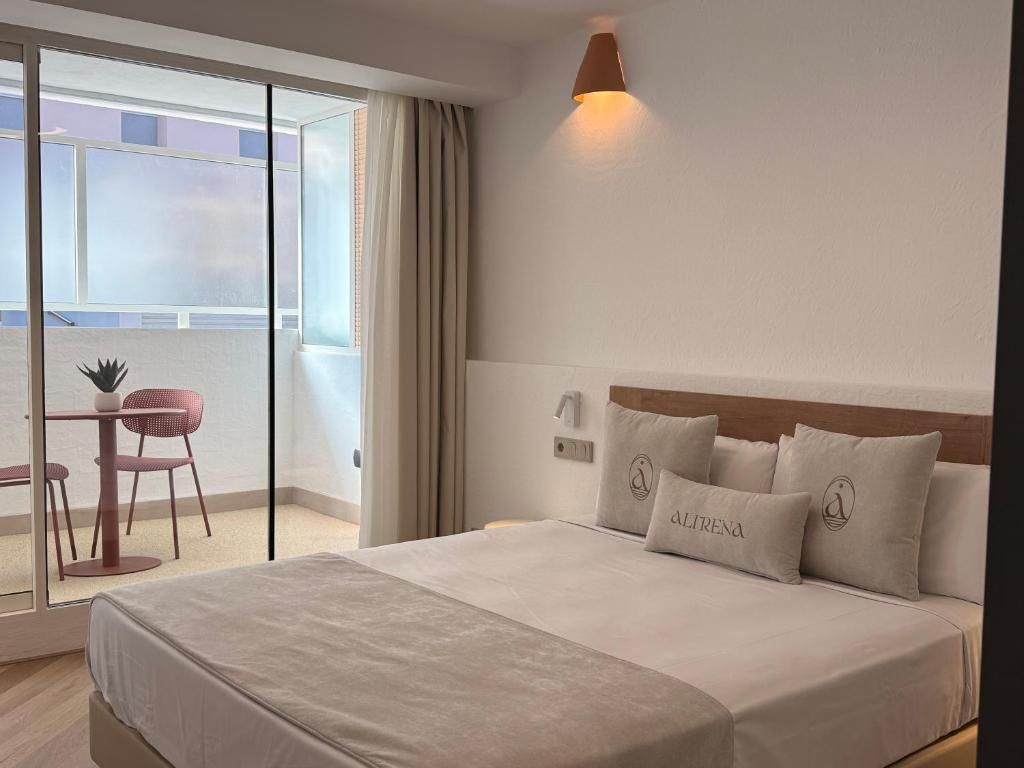 Et hotelværelse med en stor seng, et natbord, en stol, et vindue med gardiner og en balkon med bord og stole.