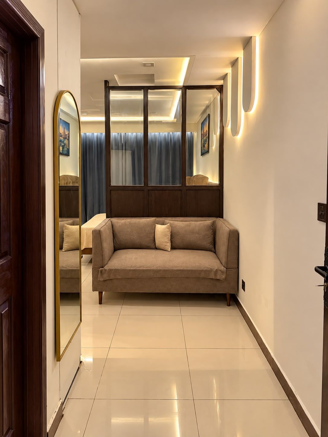 A modern hotel room with a large sofa, a mirror, and a view of a cityscape through the window.