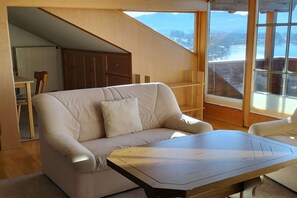 A modern living room with a sofa, a wooden coffee table, and a view of a lake through large windows. - Holiday apartment with 1 bedroom (Hopfen am See)