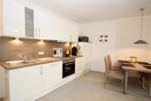 A modern kitchen with white cabinets, a built-in oven, a coffee maker, and a dining area with a table and chairs. - Holiday apartment with 2 bedrooms (Norddeich)