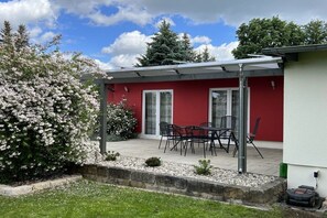 Eine Terrasse mit Tisch und Stühlen, ein rotes Gebäude mit Glastüren und ein überdachter Sitzbereich.