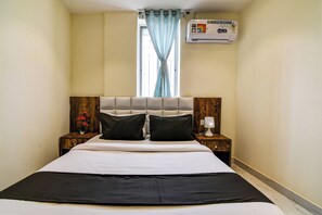 Een hotelkamer met een bed, een nachtkastje met een lamp, een raam met gordijnen en een airconditioning op de muur.