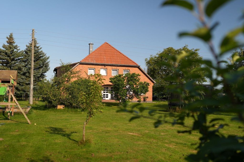 Une maison à deux étages, construite en briques et surmontée d’un toit rouge, entourée d’une cour verdoyante et d’arbres.