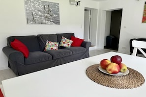 A living room with a grey sofa, red and patterned cushions, a white table with a woven placemat, and a plate of apples. - Holiday apartment with 2 bedrooms (Preetz)