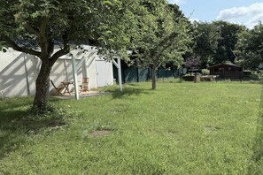 Une cour arrière verdoyante avec un arbre, un banc en bois et un bâtiment blanc doté d’un porche.