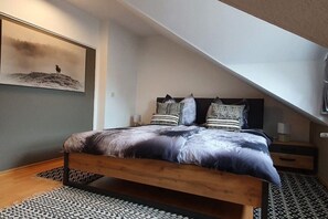 Ein Schlafzimmer mit schräg zulaufender Decke, einem Bett mit gemusterten Textilien, einem Nachttisch und einem Wandbild.