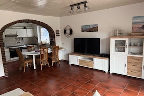 A kitchen with white cabinets, a dining table with chairs, a TV, and a cabinet with glass doors. - Holiday home with 3 bedrooms (Wangerland)