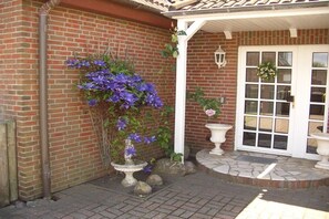 A brick building with a white door, a white porch, and a flower planter with purple flowers. - Holiday apartment with 1 bedroom (Westerland)