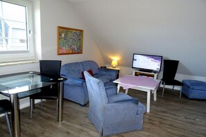 A living room with a glass table, blue sofa, a TV, and a painting on the wall. - Holiday apartment with 1 bedroom (Westerland)