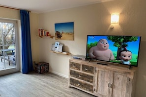 Ein Wohnzimmer mit Fernseher, Holzschrank, Bücherregal und einem Fenster mit Blick ins Freie.