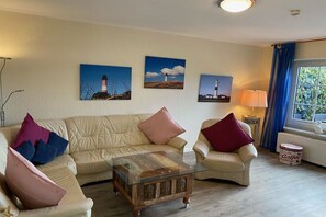 Ein Wohnzimmer mit einem beigefarbenen Sofa, einem Glastisch und einer Lampe. An der Wand hängen drei Gemälde.