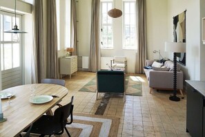 A well-lit room with a dining table, chairs, a sofa, and a lamp. - Duinweg 4a - Apartment A (Huisduinen)
