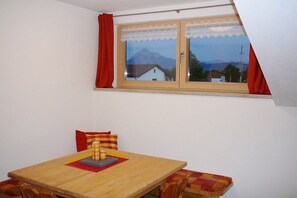 A room with a wooden table and bench, a window with curtains, and a view of houses and mountains. - At the bricklayer's - Alpine rose (Hopferau)