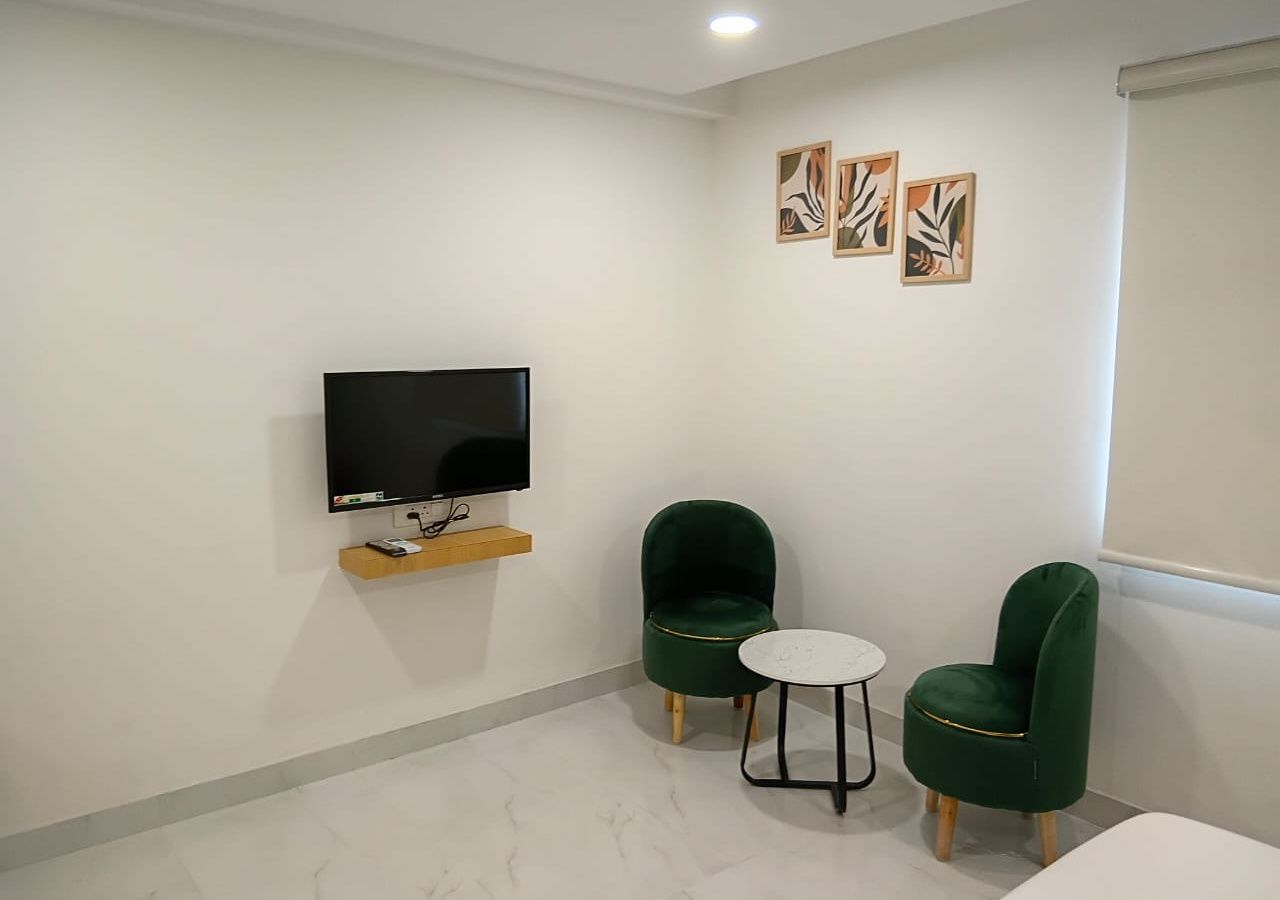 Une pièce avec un téléviseur à écran plat fixé au mur, deux fauteuils verts, une petite table ronde et une étagère en bois située sous le téléviseur.
