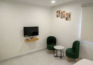 A room with a flat-screen TV mounted on the wall, two green upholstered chairs, a small round table, and a wooden shelf below the TV. - Hotel Prince Inn (Chandrapur)