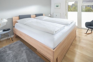 A double bed with a wooden frame and white bedding in a room with a grey rug, a small table with a lamp, and a chair. - Holiday apartment in Juist with swimming pool (Juist)