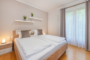 A bedroom with a wooden bed, white bedding, a bedside table with a lamp, and a window with curtains. - Delbrück-Villen D125 Vacation Rental on the Beachfront in Heringsdorf (Heringsdorf (Seebad))
