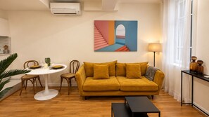 Un salon avec un canapé jaune, une table à manger ronde et un tableau accroché au mur.