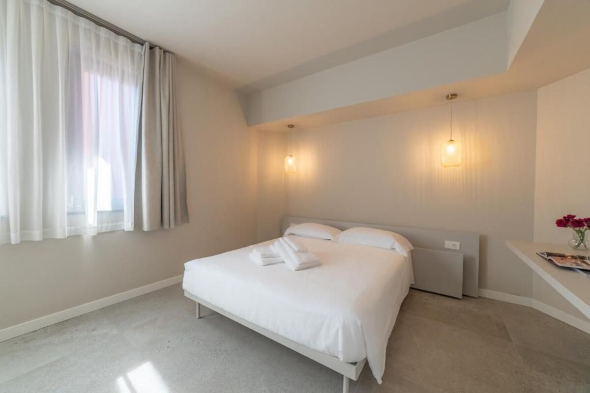 Un dormitorio con cama, mesita de noche, ventana con cortinas e iluminación en la pared.
