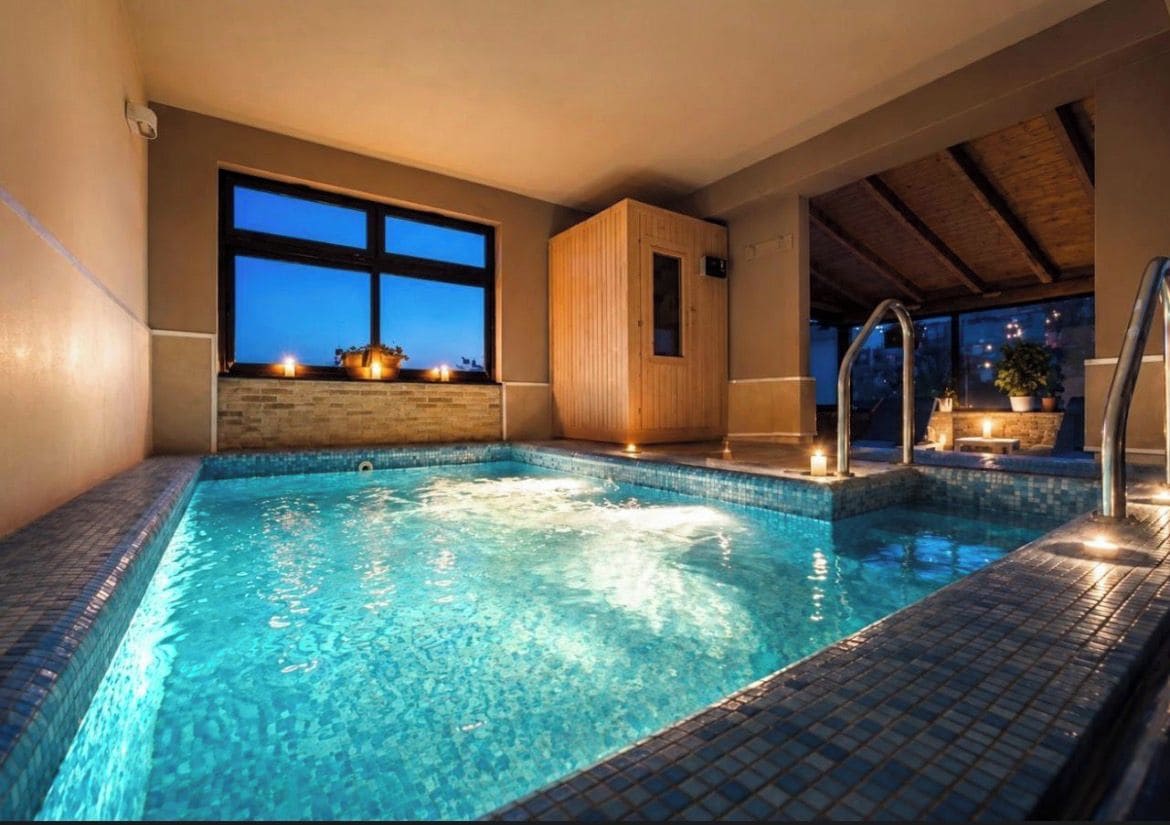 A heated indoor swimming pool with a wooden sauna, a stone wall, and a view of the evening sky.