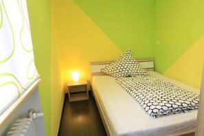 Ein kleines Zimmer mit einem Einzelbett, einem Nachttisch, einer Lampe und einem Fenster mit Vorhängen.