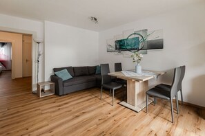 A modern living room with a wooden floor, a grey sofa with teal cushions, a dining table with chairs, and a wall-mounted abstract art piece. - Holiday apartment with 2 bedrooms (Fischen im Allgäu)