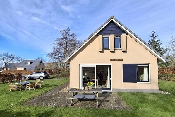 Een gelijkvloers huis met schuin dak, een voordeur en een klein terras met een tafel en stoelen.