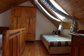 A bedroom with a sloped ceiling, a wooden wardrobe, a bed with a plaid bedspread, and a window with a plaid curtain. - Gutmann's cellar house (Fehring)