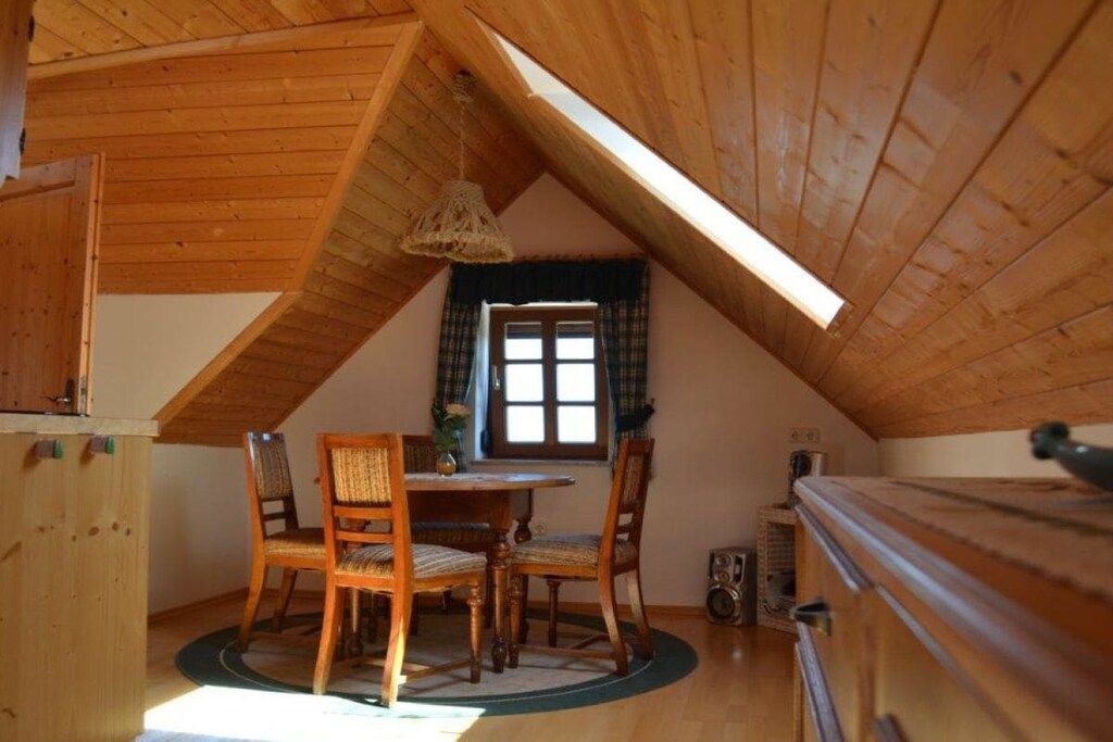 A cozy attic room with a sloped ceiling, wooden furniture, a dining table, and a window with plaid curtains.