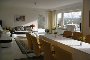 Un salon moderne avec un canapé, une table à manger et des chaises, et une vue sur l’extérieur grâce à de grandes fenêtres.