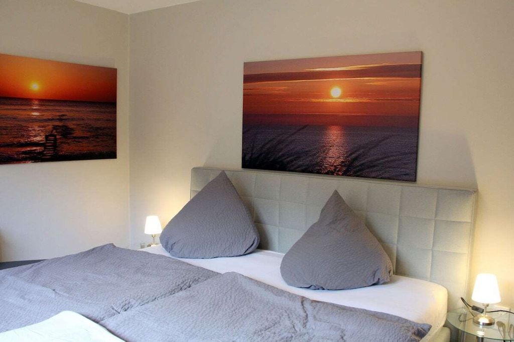 A hotel room with a bed, two pillows, a headboard, and two wall-mounted paintings of a sunset over the ocean.