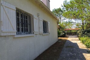 A building with white walls and shutters, a paved pathway, and a fenced area with trees. - "Le Bateau" by the sea (Vandays Montalivet)