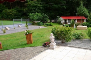 A courtyard with a statue, a small building with a red roof, and a fenced sports field. - Holiday home in Mehren with swimming pool (Mehren)