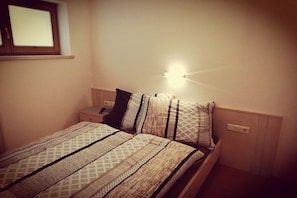 Een slaapkamer met een bed, kussens, een nachtkastje, een stopcontact en een raam.