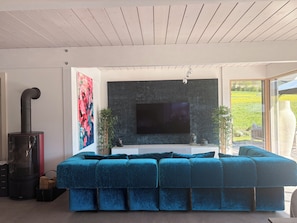 A modern living room with a sectional sofa, a flat-screen TV mounted on a textured wall, and a large window offering a view of a garden. - Architect-designed home with a heated pool, just a stone's throw from Geneva and the ski slopes (Présilly)